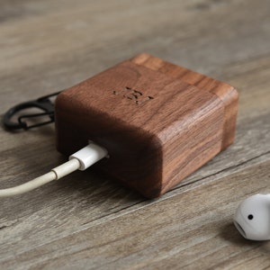 May include: A wooden charging dock for wireless earbuds, with a white charging cable plugged in. The dock is made of dark wood and has a rounded shape. A white earbud is lying on the wooden surface next to the dock.