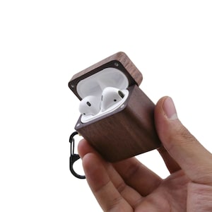 May include: A brown wooden case with a hinged lid, open to reveal two white wireless earbuds. The case has a black metal carabiner attached.