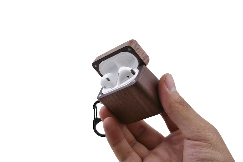 May include: A brown wooden case with a hinged lid, open to reveal two white wireless earbuds. The case has a black metal carabiner attached.