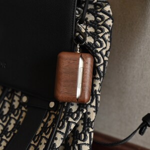 May include: A brown wooden case with a silver metal clasp attached to a black and white patterned bag with a black strap. The case is hanging from the bag with a silver metal carabiner.
