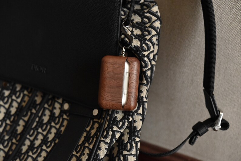 May include: A brown wooden case with a silver metal clasp attached to a black and white patterned bag with a black strap. The case is hanging from the bag with a silver metal carabiner.