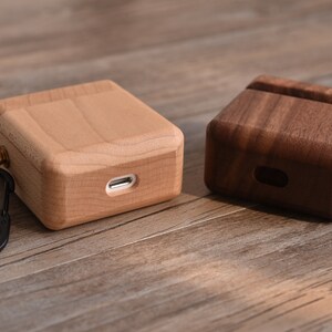 May include: Two wooden cases for wireless earbuds, one made of light wood and the other of dark wood. Both cases have a small metal loop with a carabiner attached.