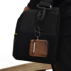 May include: A brown wooden case with a silver accent hangs from a black carabiner attached to a black bag. The case is attached to a black and yellow strap.