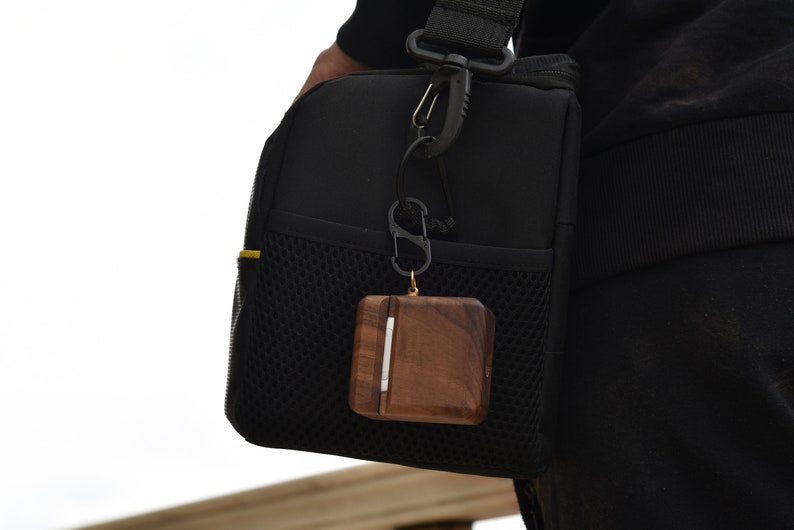 May include: A brown wooden case with a silver accent hangs from a black carabiner attached to a black bag. The case is attached to a black and yellow strap.