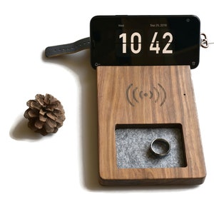 May include: A wooden charging station with a grey felt-lined compartment for a ring, a wireless charging pad, and a phone stand. The phone stand has a black leather strap and a black phone with a white digital clock displaying "10:42".