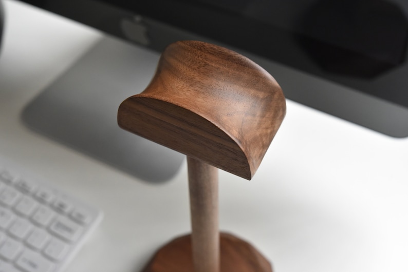 May include: A wooden headphone stand with a curved top and a round base. The stand is made of dark brown wood.