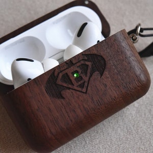 May include: A dark brown wooden case for wireless earbuds with a Superman logo engraved on the front. The case has a metal carabiner attached to it.