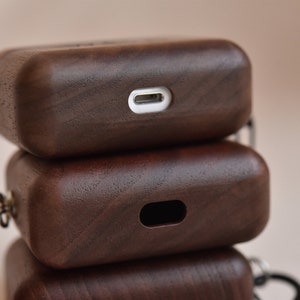 May include: Three stacked wooden cases for wireless earbuds, each with a different design. The top case has a charging port, the middle case has a cutout for a charging port, and the bottom case has a metal loop.
