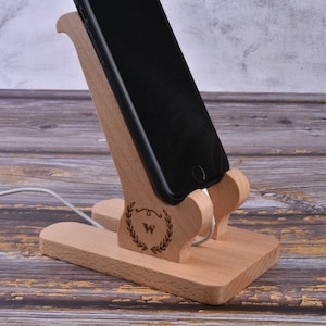 Op de afbeelding: Houten telefoonstandaard met een gesneden ontwerp en de letter "W" in een krans. De standaard is ontworpen om een telefoon rechtop te houden en heeft een gleuf voor oplaadkabels.