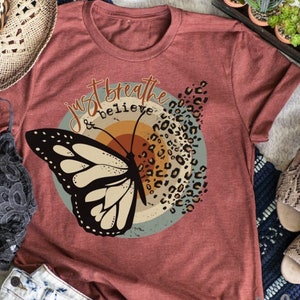 May include: A maroon t-shirt with a graphic of a butterfly with a sunset background and the text "Just Breathe & Believe".