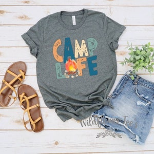 May include: A gray t-shirt with the text "Camp Life" in colorful, distressed letters. A campfire with flames is in the center of the text.
