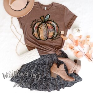 May include: A brown t-shirt with a tie-dye pumpkin graphic. The shirt is paired with a black and white polka dot skirt, a white purse, and brown ankle boots.