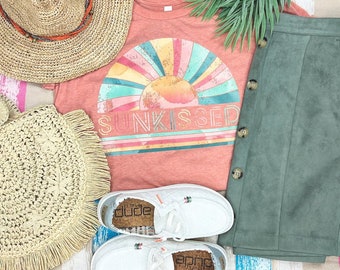Può includere: Una maglietta color corallo con una grafica di tramonto retrò e il testo "Sunkissed". La maglietta è abbinata a una gonna di camoscio verde, un cappello di paglia, una borsa di paglia e scarpe di tela bianche con suola in sughero.