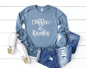 May include: A casual outfit featuring a blue long-sleeve shirt with the words "coffee & books" in white script, paired with denim shorts and white sneakers. The ensemble is laid out on a white wooden surface.