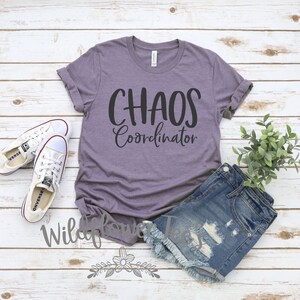 May include: A purple t-shirt with the words "CHAOS Coordinator" in black script. The shirt is paired with denim shorts, white sneakers, and a small plant. The image is set on a white wooden background.
