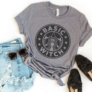 May include: A gray t-shirt with a black and white graphic of a witch with a witch hat in a circle with the text "BASIC WITCH" around the circle. The shirt is paired with denim shorts and black shoes.