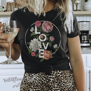 May include: Black t-shirt with a white circle graphic featuring the word "LOVE" surrounded by pink, white, and green floral illustrations.