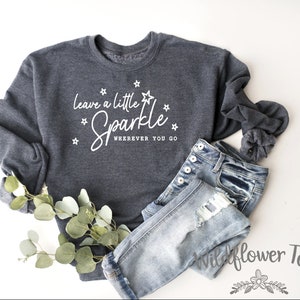 May include: A dark gray sweatshirt with the text "Leave a little sparkle wherever you go" in white lettering. The sweatshirt is decorated with small white stars.