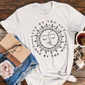 May include: A light gray t-shirt with a circular graphic featuring a sun and moon design, with the text "LIVE BY THE SUN LOVE BY THE MOON". The shirt is styled with denim shorts, glasses, a book, and a cup of tea.