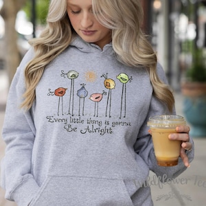 May include: A light gray hoodie with a front pocket and a drawstring hood. The hoodie features a whimsical design of colorful birds on long stems, with the text "Every little thing is gonna Be Alright".