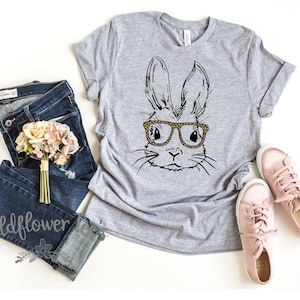 May include: A gray t-shirt with a black and white illustration of a bunny wearing leopard print glasses. The shirt is laid flat on a white surface with a pair of pink sneakers, a bouquet of pink flowers, and a pair of blue jeans.