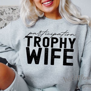 May include: A light gray sweatshirt with the words "participation" above and "TROPHY WIFE" in large, bold, black letters. The sweatshirt has a crew neck and long sleeves. The brand name "Wildflower Tees" is printed on the sleeve.