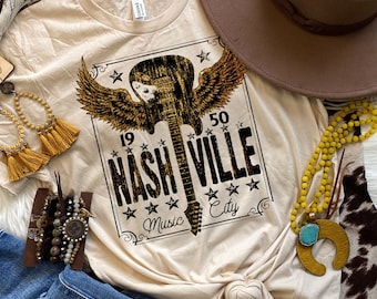 May include: Cream-colored t-shirt featuring a distressed graphic of a guitar with wings, the text "NASHVILLE" and "Music City". The shirt is styled with a brown hat, jewelry, and denim.