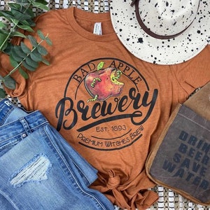 May include: A rust-colored t-shirt with a graphic of a red apple with a bite taken out of it. The graphic says "Bad Apple Brewery Est. 1693 Premium Witches Brew".