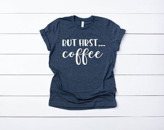 May include: A navy blue t-shirt with the white text "BUT FIRST... coffee". The shirt is made of a soft material and is displayed on a white wooden surface. The sleeves are cuffed.