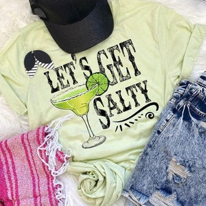 May include: A light green t-shirt with the text "Let's Get Salty" and a margarita glass with a lime wedge. The shirt is laid out on a white fuzzy surface with a pink and white striped blanket, a black baseball cap, and blue denim shorts.