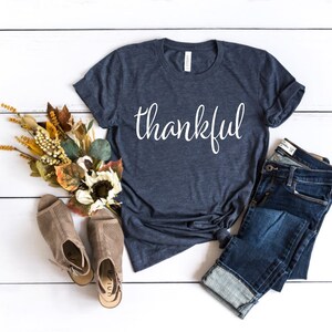 May include: A navy blue t-shirt with the word "thankful" in white script. The shirt is paired with folded blue jeans, tan open-toe booties, and a fall-themed floral arrangement with sunflowers and dried leaves.