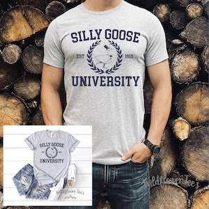 May include: A light gray t-shirt with the words "SILLY GOOSE UNIVERSITY" in navy blue. The design includes a goose wearing a top hat inside a laurel wreath. The shirt is displayed on a person wearing blue jeans.