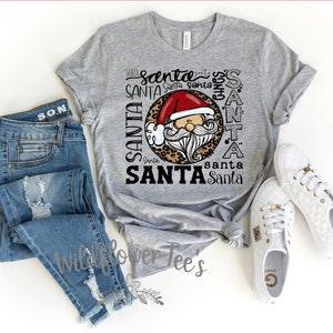 May include: A heather gray t-shirt featuring a Santa Claus graphic with a red hat and white beard. The word "Santa" is repeated in various fonts around the Santa image. The shirt is paired with blue jeans and white sneakers.