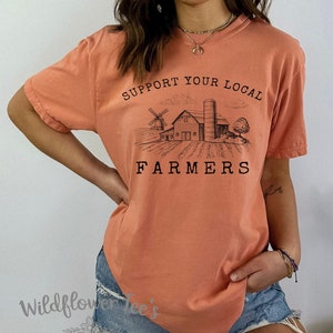 K&ouml;nnte beinhalten: Ein pfirsichfarbenes T-Shirt mit einer Grafik einer Farmszene und dem Text "Unterst&uuml;tze deine lokalen Bauern".