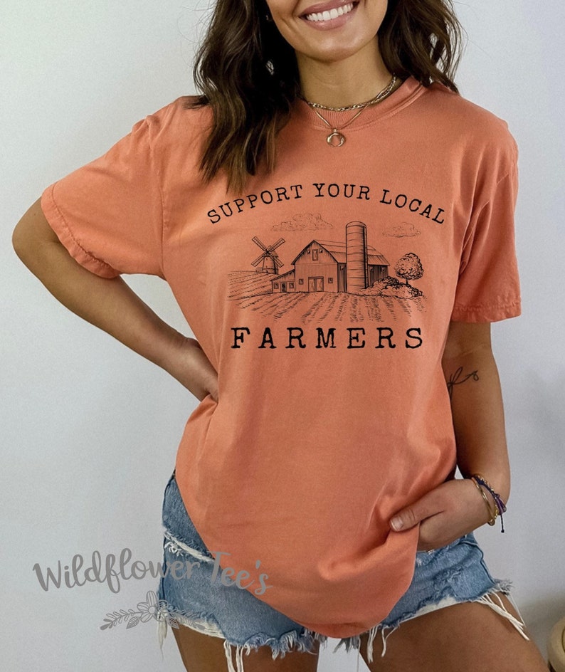 K&ouml;nnte beinhalten: Ein pfirsichfarbenes T-Shirt mit einer Grafik einer Farmszene und dem Text "Unterst&uuml;tze deine lokalen Bauern".