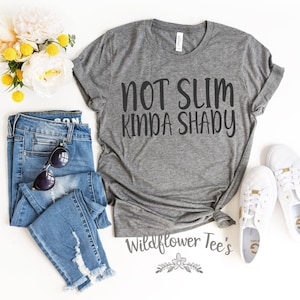 May include: Gray t-shirt with the text "Not Slim Kinda Shady" in black lettering. The shirt is displayed on a white background with a pair of jeans and white sneakers.
