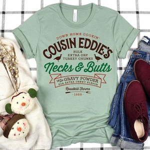 May include: A light green t-shirt with the text "COUSIN EDDIE'S" in brown and green, featuring turkey illustrations. The shirt also includes the phrases "Necks & Butts" and "Gravy Powder". The text is in a vintage style.