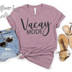 May include: A mauve t-shirt with the words "Vacay Mode" in black script. The shirt is paired with denim shorts, sunglasses, and brown sandals with gold accents. The Wildflower Tee's logo is in the upper left corner.