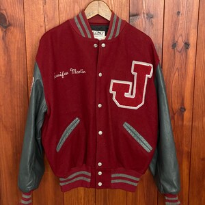 Op de afbeelding: Een bordeauxrode en grijze varsity-jas met grijze leren mouwen. De jas heeft een groot wit en rood "J"-embleem en de naam "Jennifer Martin" op de voorkant geborduurd. Grijze en witte gestreepte biezen accentueren de kraag, manchetten en taille.