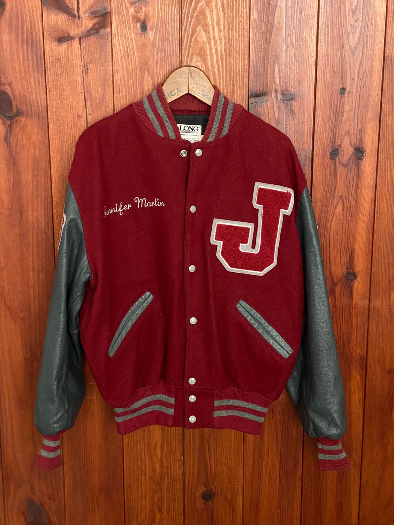 Op de afbeelding: Een bordeauxrode en grijze varsity-jas met grijze leren mouwen. De jas heeft een groot wit en rood "J"-embleem en de naam "Jennifer Martin" op de voorkant geborduurd. Grijze en witte gestreepte biezen accentueren de kraag, manchetten en taille.