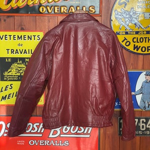 May include: A burgundy leather jacket with a bomber-style design, featuring a ribbed hem and cuffs. The jacket has a classic collar and is hanging on a wooden hanger. Vintage-style signs are in the background.