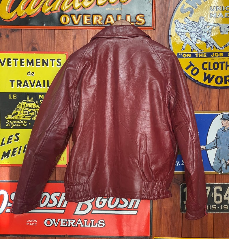 May include: A burgundy leather jacket with a bomber-style design, featuring a ribbed hem and cuffs. The jacket has a classic collar and is hanging on a wooden hanger. Vintage-style signs are in the background.