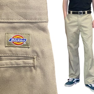 May include: A pair of khaki Dickies pants with a back pocket and a small Dickies logo patch. The pants are worn by a person standing with their hands in their pockets.