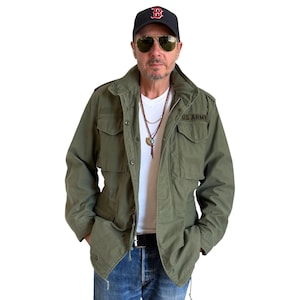 May include: An olive green US Army jacket with a white t-shirt. The jacket has two chest pockets and the words "US ARMY" printed on the right chest. The person is wearing a black baseball cap and aviator sunglasses.