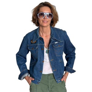 May include: A classic denim jacket in a medium blue wash, with a button-up front and chest pockets. The jacket has a collar and a small patch on the upper left chest. The model is wearing white sunglasses and green trousers.