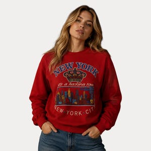 Op de afbeelding: Rode sweatshirt met de tekst "NEW YORK" in blauw boven een kroongrafiek. Onder de kroon staat de zin "it's a hecking town" en een skyline-afbeelding. De woorden "NEW YORK CITY" staan onderaan.
