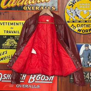 May include: A burgundy leather jacket with a red quilted lining. The jacket has a collared neckline, snap closures, and a ribbed hem. The interior lining is visible, and the jacket is displayed against a backdrop of vintage signs.