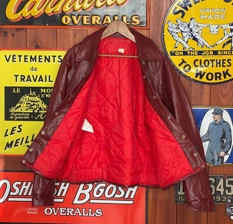 May include: A burgundy leather jacket with a red quilted lining. The jacket has a collared neckline, snap closures, and a ribbed hem. The interior lining is visible, and the jacket is displayed against a backdrop of vintage signs.