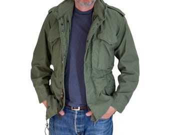 May include: An olive green field jacket featuring a high collar, shoulder epaulets, and four front pockets with snap-button flaps. The jacket is worn over a dark gray t-shirt and blue denim jeans, with a visible metal zipper and waist drawstring.