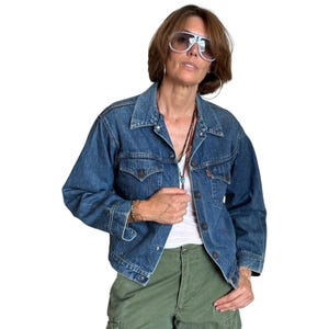 May include: A vintage denim jacket in a medium wash, featuring a classic collar, button-up front, and two chest pockets. The jacket has a relaxed fit and is paired with green cargo pants. The model is wearing sunglasses and a necklace.